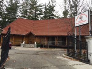 Photo taken at the front gates of the Buddhist Centre Toronto Mahavihara which shows the main building with a red roof and brown walls. The front steps are visible to the main door and a ramp on the left.